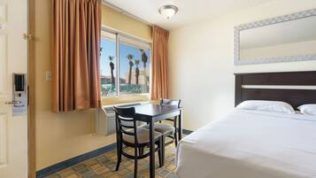 Una habitación de hotel con cama, una mesa y dos sillas, un ventanal con cortinas y vista a palmeras.