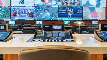 Una sala de control con múltiples monitores mostrando diversos eventos deportivos, un panel de control y una zona de asientos.