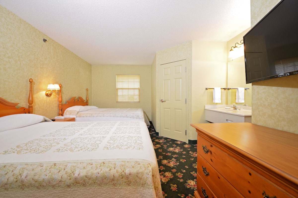 A hotel room with two beds, a wooden dresser, a flat-screen TV, and a bathroom with a mirror and towel rack.