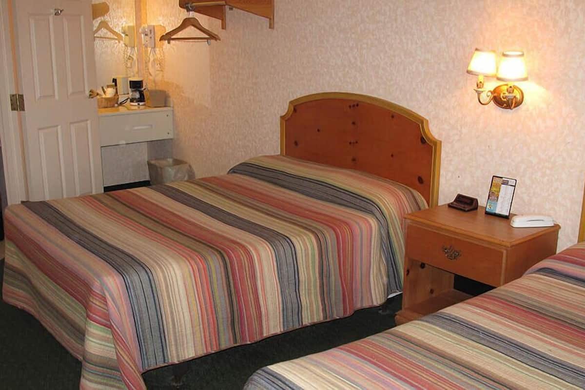 A hotel room with two beds, a wooden headboard, a bedside table with a lamp, a small desk with a phone and a magazine, and a wall-mounted light fixture.