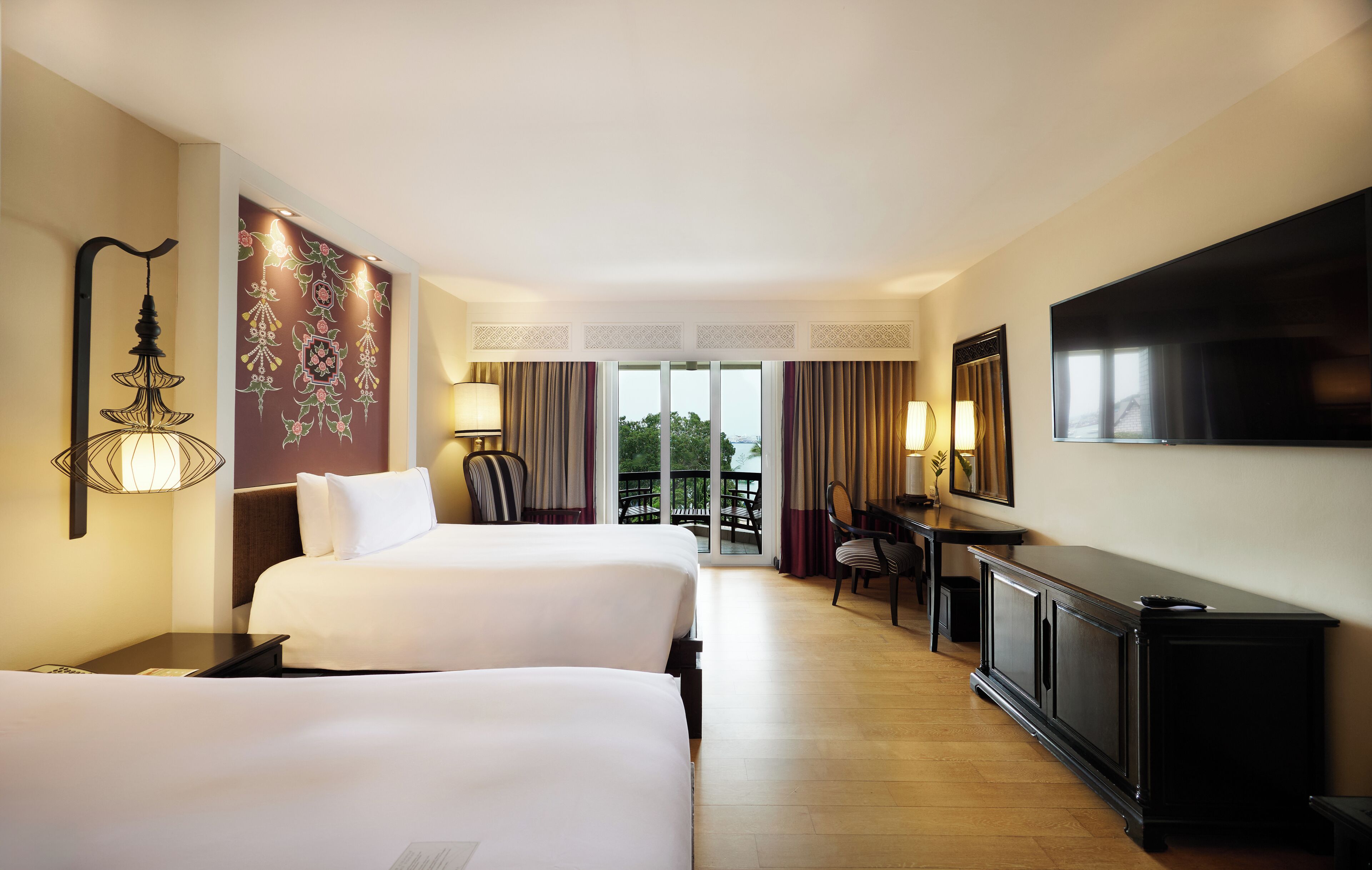 A hotel room with two beds, a desk, a TV, and a balcony with curtains.