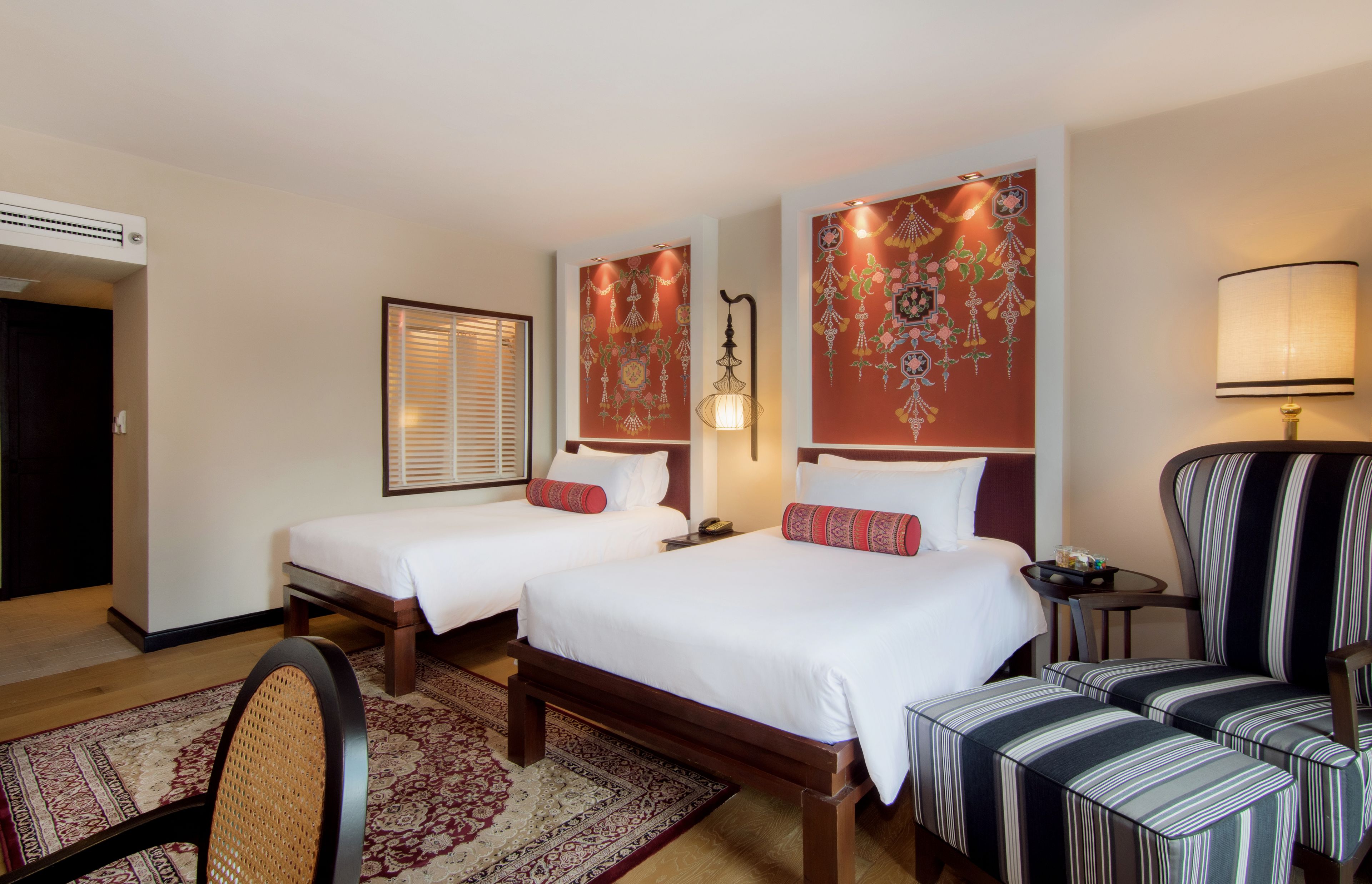 A hotel room with two beds, a sitting area with a chair, a lamp, and a decorative wall hanging.