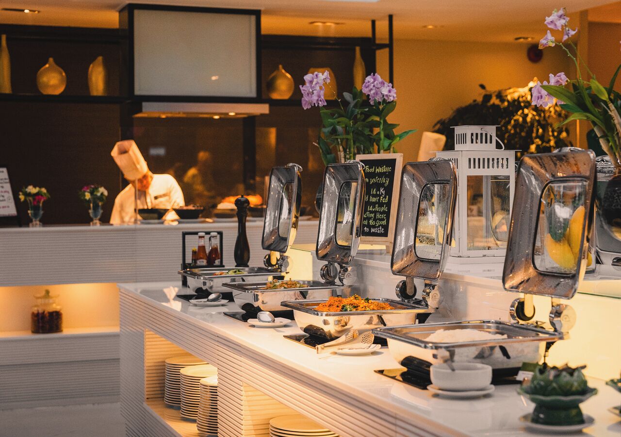 A buffet setup with multiple food stations, a chef in the background, and decorative elements like flowers and vases.