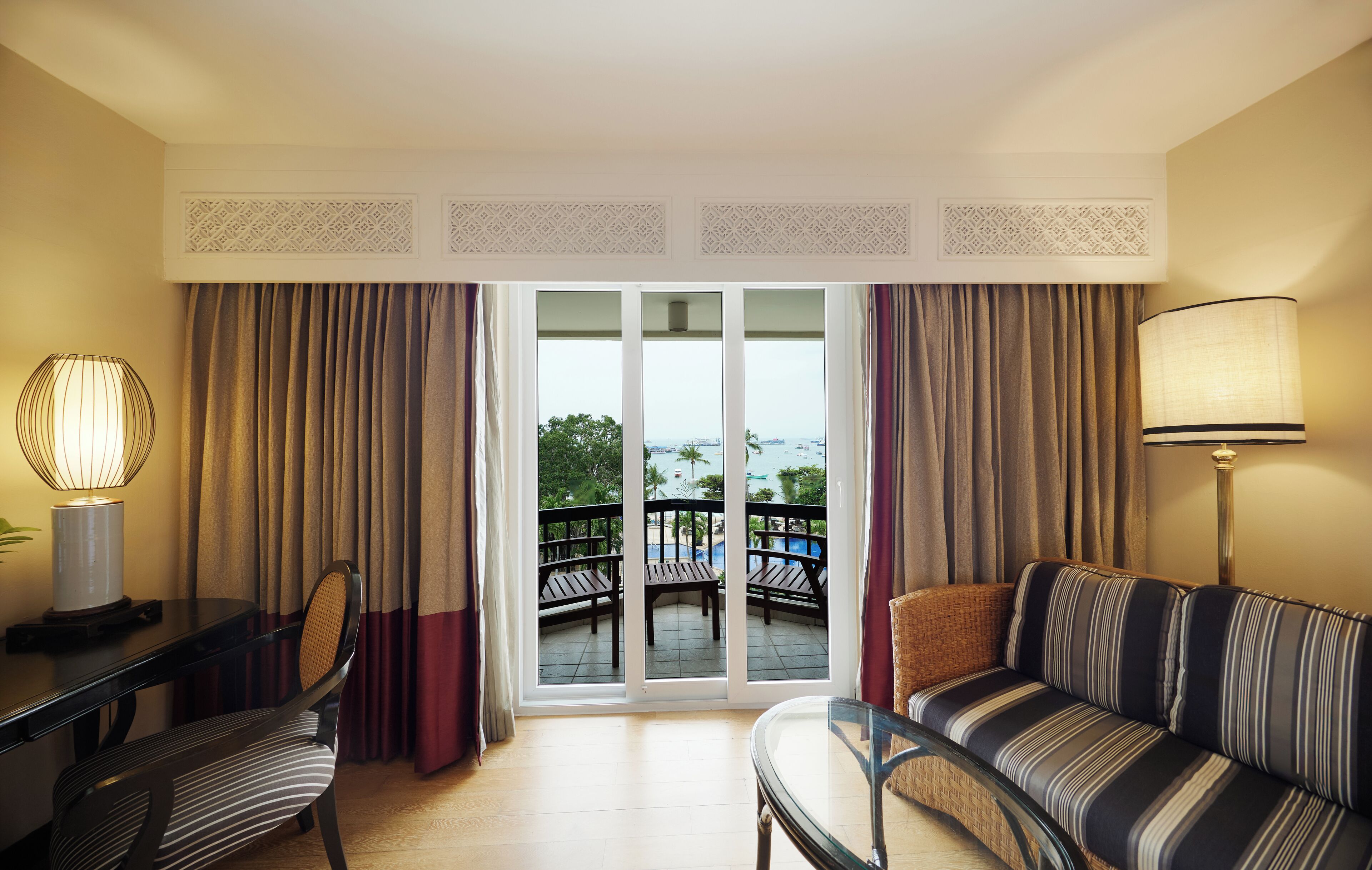 A hotel room with a balcony, a sofa, a dining table with chairs, and a lamp.