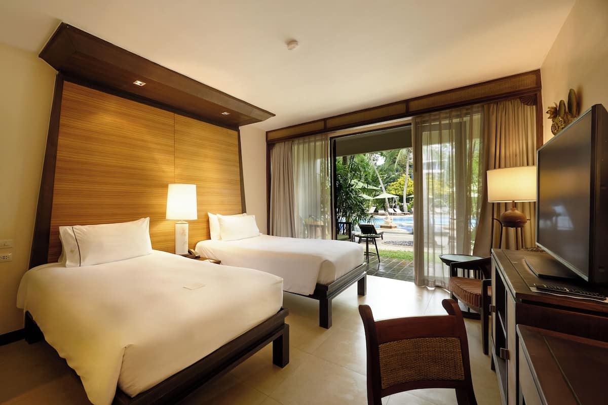 A hotel room with two beds, a TV, a desk, and a view of a pool and greenery.
