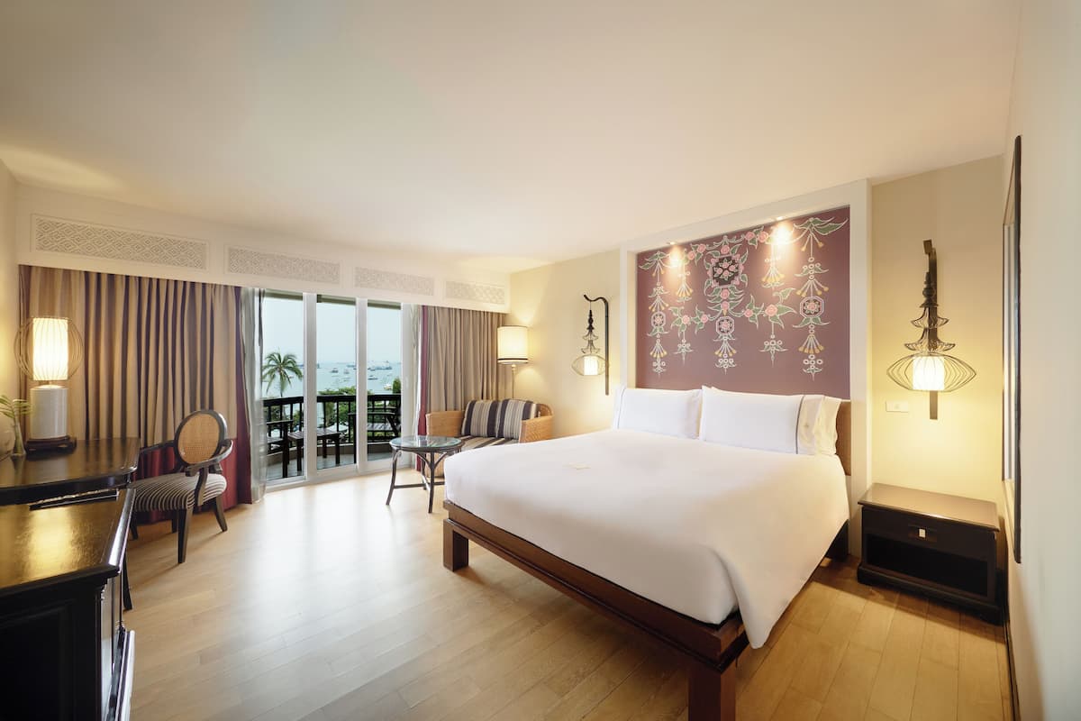 A hotel room with a large bed, a desk, a chair, a small table, and a balcony with a view.