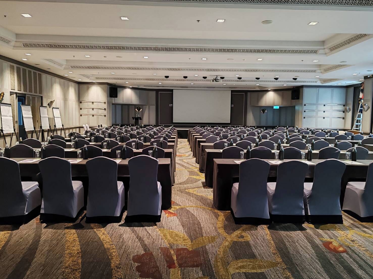 Un salón de conferencias con filas de mesas y sillas, una pantalla de proyector y suelo alfombrado.