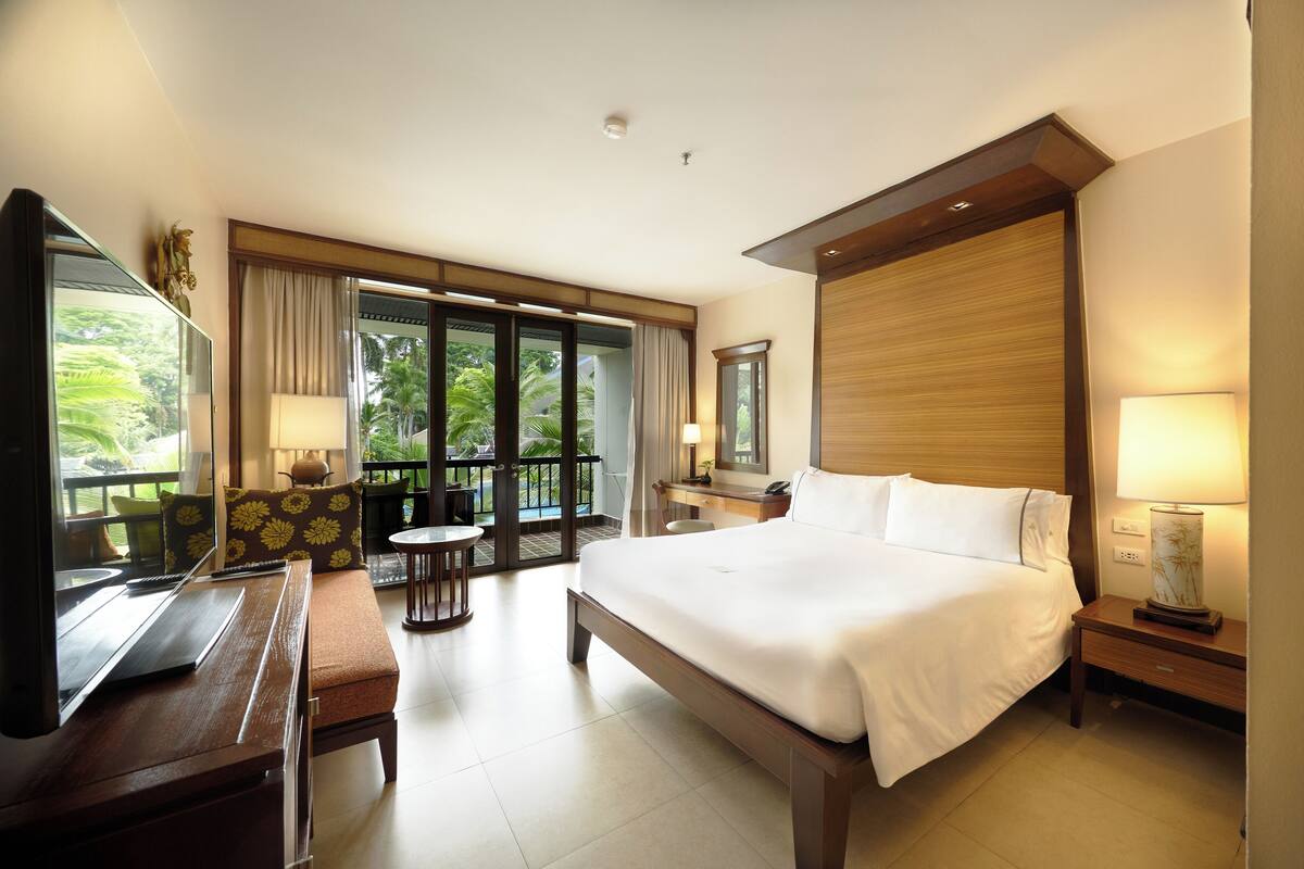 A hotel room with a large bed, a desk with a laptop, a TV, and a balcony with a view of palm trees.