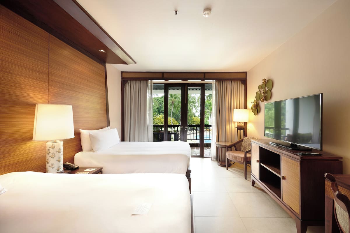 A hotel room with two beds, a TV, a balcony with a view of greenery, and a wooden wall.