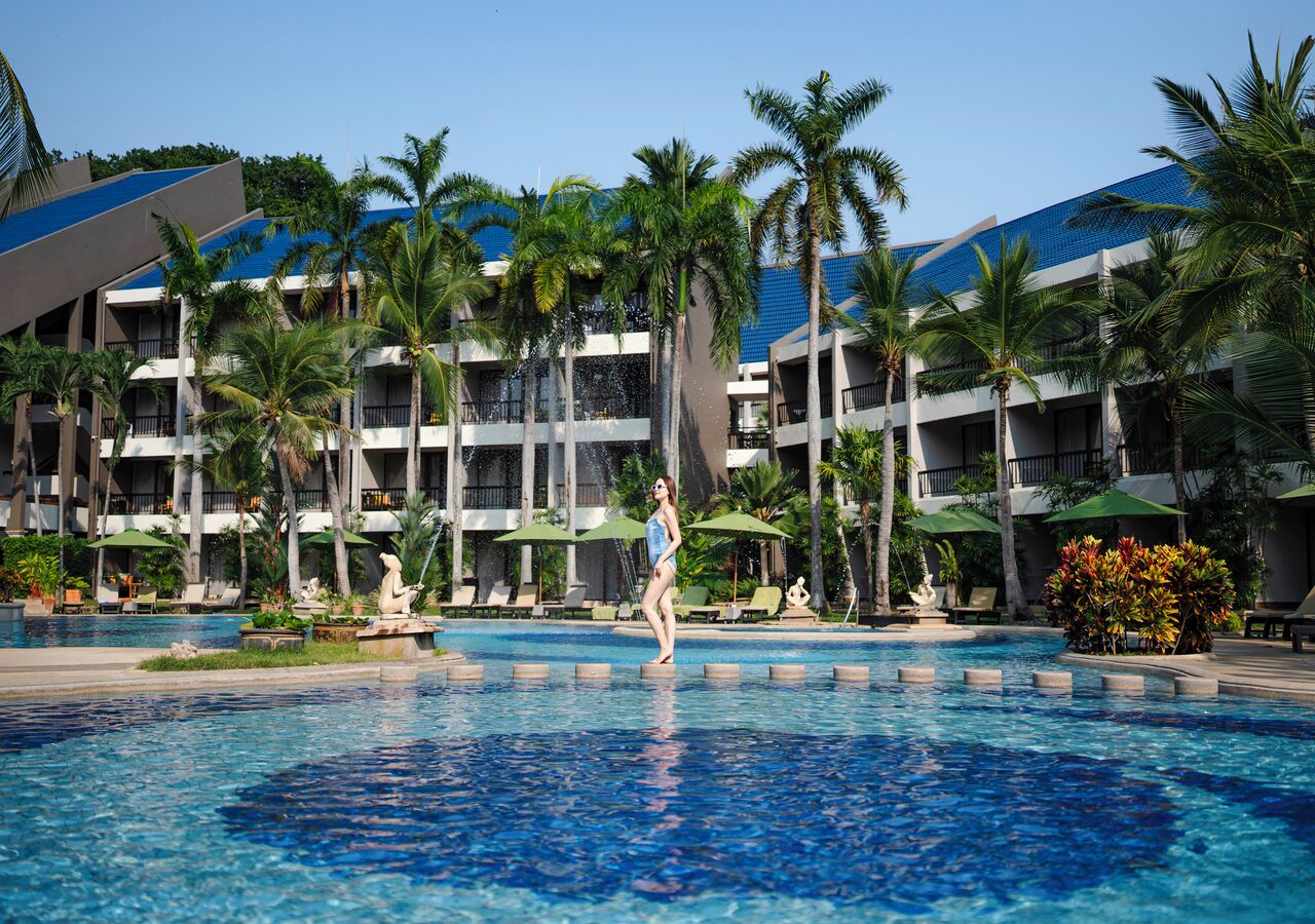 Piscina de un resort con palmeras, sillas de descanso y un edificio con balcones al fondo.