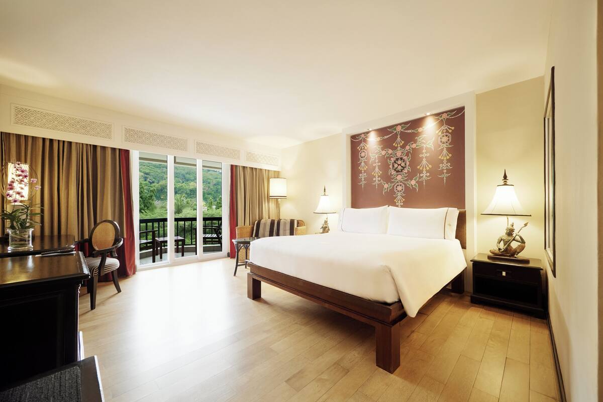 A hotel room with a large bed, a desk with a chair, a lamp, and a view of the outdoors.