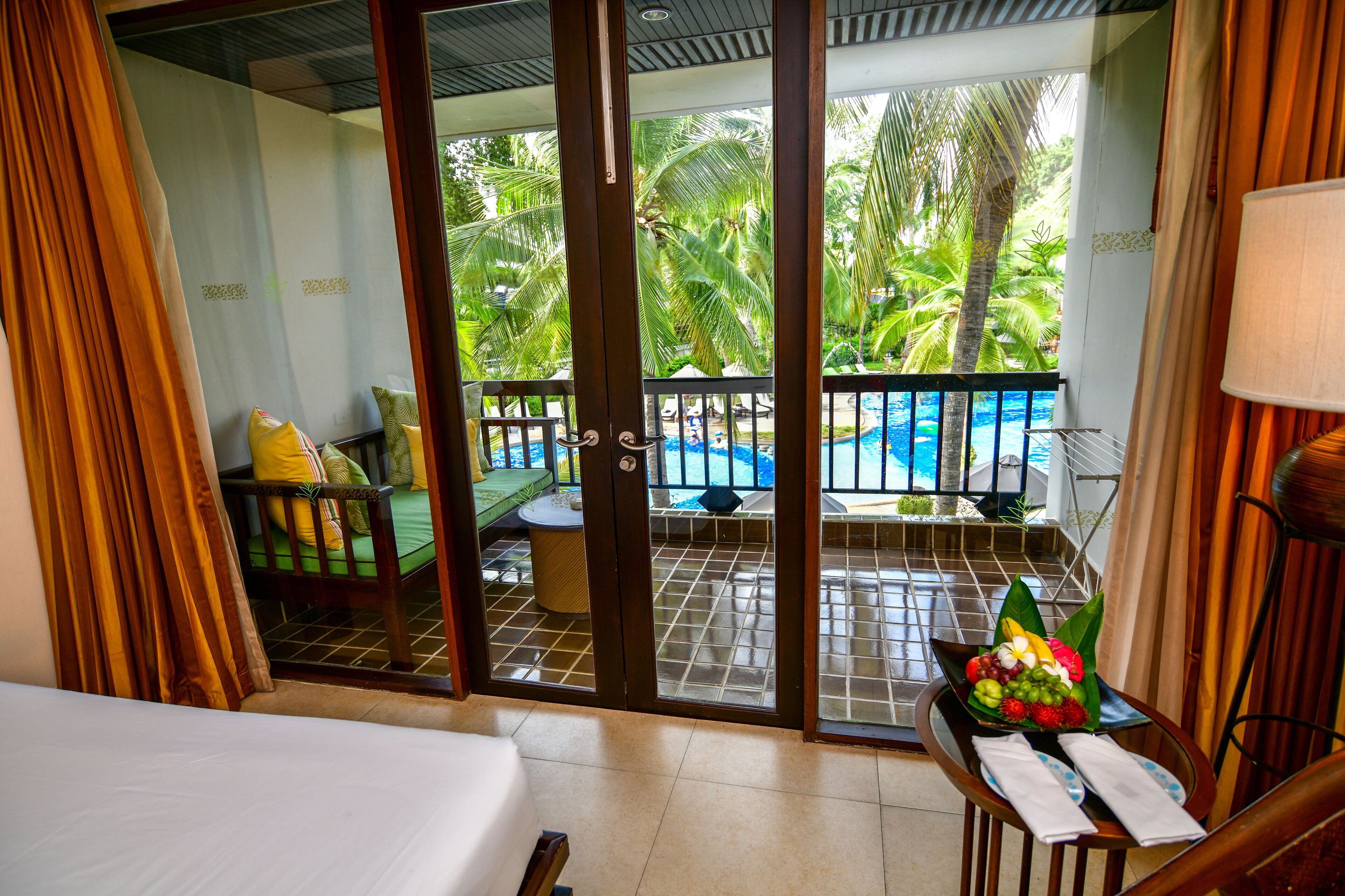 Camera d'albergo con balcone vista piscina, un letto e un tavolo con un'composizione di frutta.