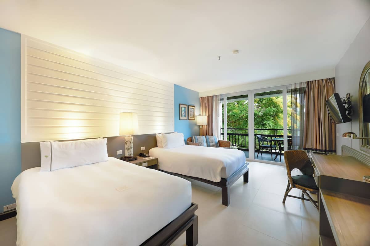 A hotel room with two beds, a desk, a chair, and a balcony with a view of greenery.