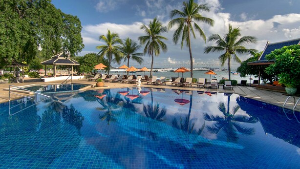 Ein Swimmingpool mit Liegestühlen und Sonnenschirmen, umgeben von Palmen und Blick auf das Meer.