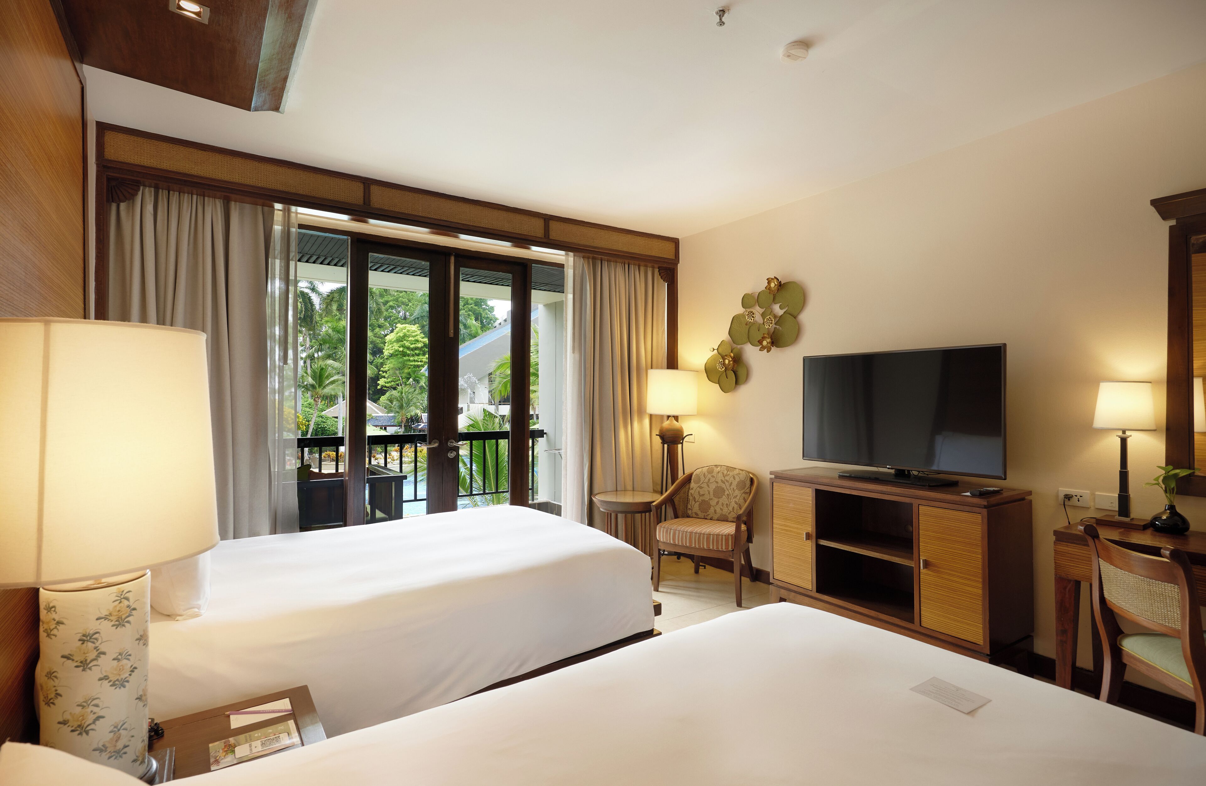 A hotel room with two beds, a television, a desk with a lamp, and a view of greenery outside.