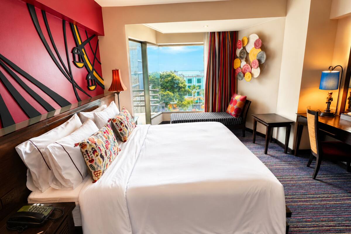 A hotel room with a bed, a desk, a chair, a telephone, a lamp, and a view of the outside.