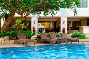 Un espace piscine avec des chaises longues, un arbre et la façade d’un bâtiment ornée de colonnes et de fenêtres.