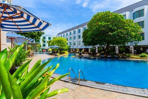 Piscina com espreguiçadeiras e guarda-sóis, cercada por um edifício com várias janelas e uma árvore.