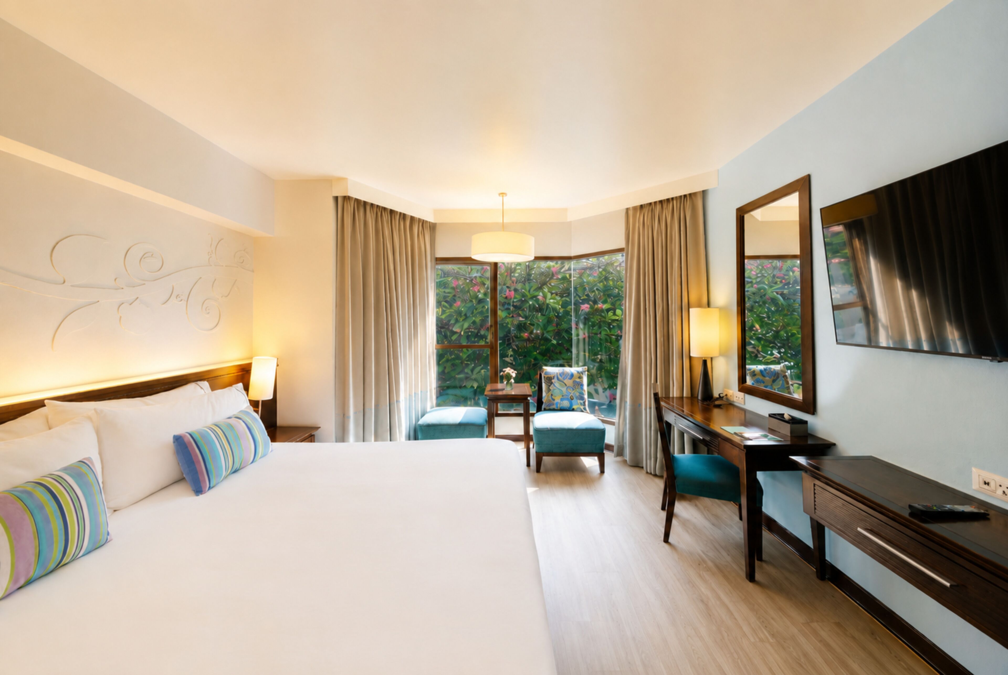 A hotel room with a large bed, a desk, a chair, a desk lamp, a bedside table, a mirror, and a window with curtains.