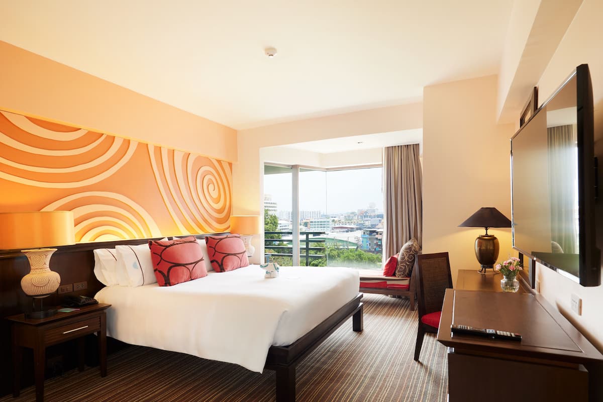 A hotel room with a large bed, a desk with a TV, a chair, a nightstand, and a view of the city through the window.