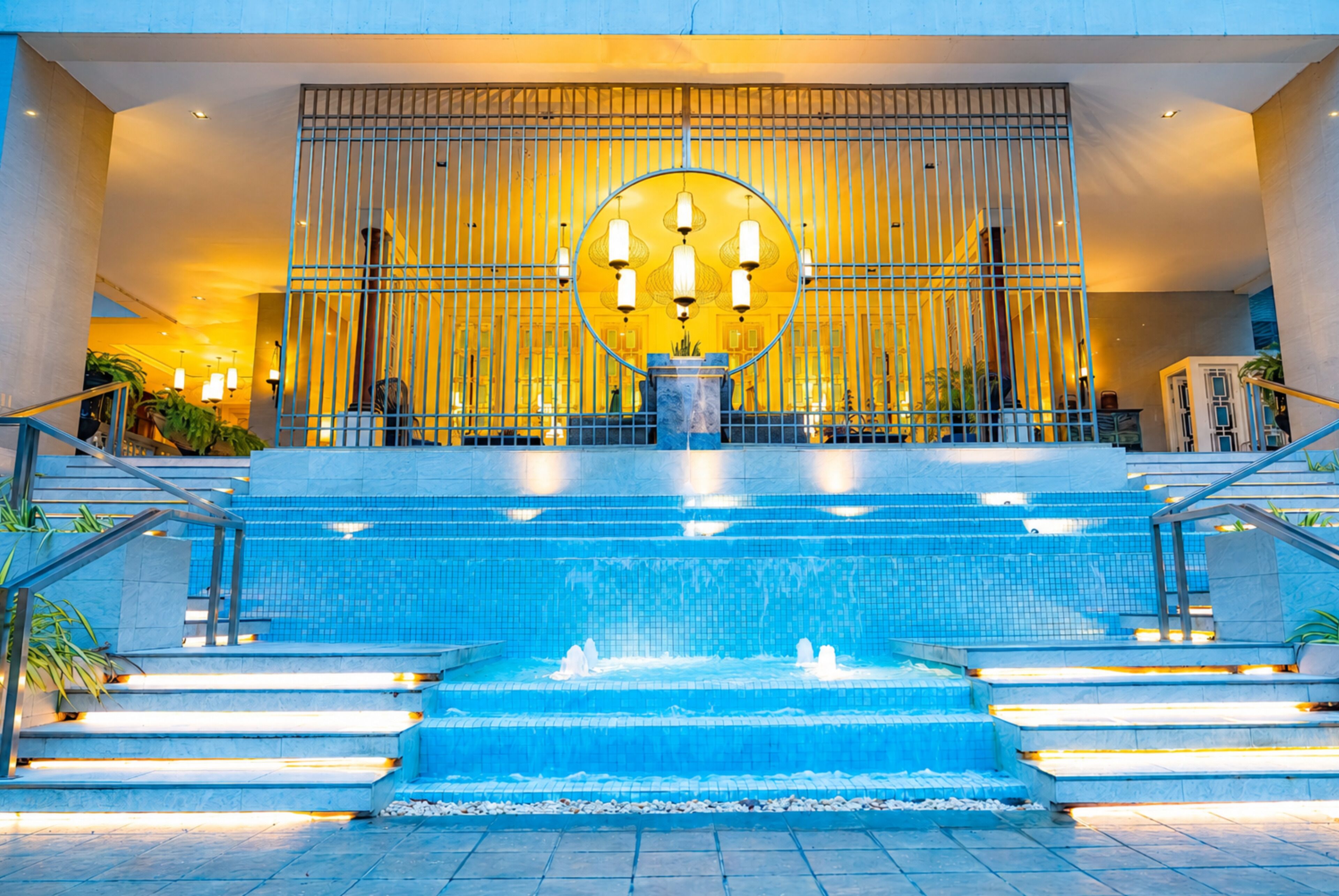 A hotel lobby with a central water feature, steps leading up to it, and a decorative wall with vertical lines.