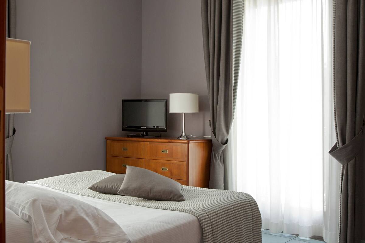 Une chambre d’hôtel avec un lit, un téléviseur posé sur une commode en bois et une fenêtre avec des rideaux.