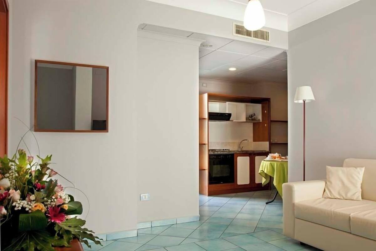 Un salon avec un canapé et une table verte, un coin cuisine avec des placards et une cuisinière, et un miroir accroché au mur.