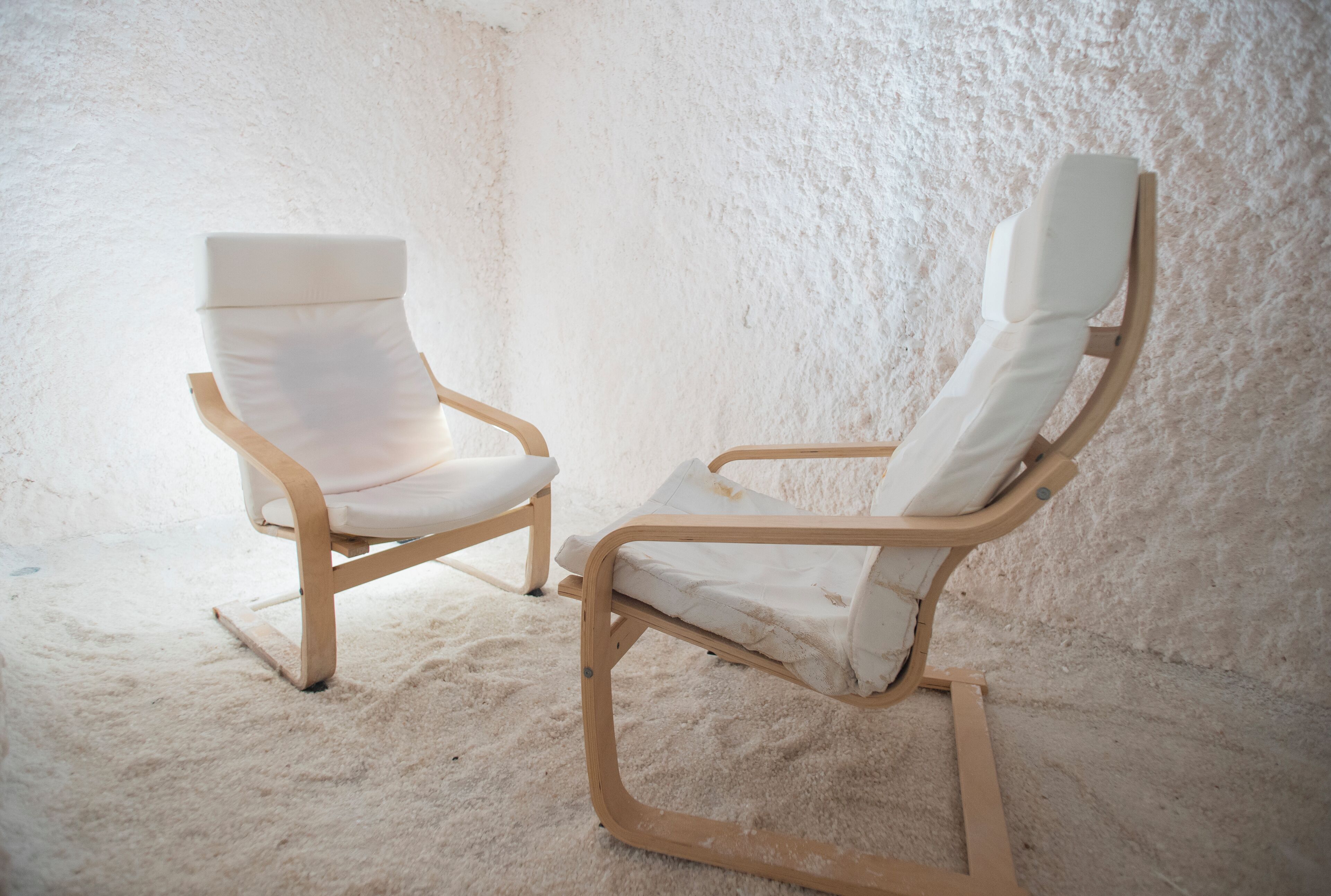 Deux fauteuils blancs à l’allure moderne, dotés d’une structure en bois, se trouvent dans une pièce dont le mur est d’un blanc texturé et le sol recouvert d’une moquette.