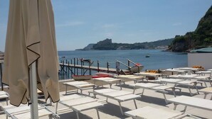 Eine Strandlounge mit Liegestühlen und Sonnenschirmen, eine Anlegestelle mit Booten und Blick auf das Meer und eine entfernte Insel.