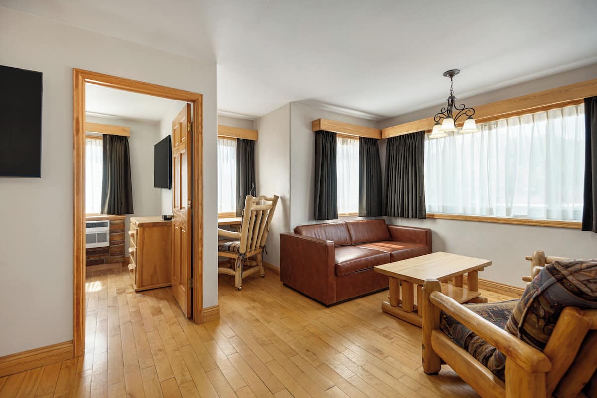 A spacious living room with a wooden floor, a brown leather sofa, a wooden coffee table, and a wooden chair. There is a television mounted on the wall, and a window with curtains.