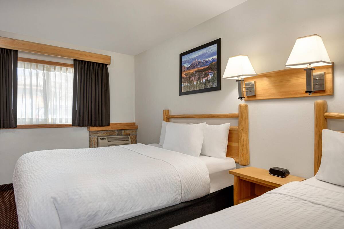 A hotel room with two beds, a wooden headboard, a nightstand, a lamp, and a painting on the wall.