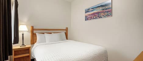 A hotel room with a bed, a nightstand with a lamp, a painting on the wall, and a window with curtains.