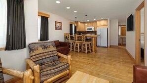 Ein geräumiges Wohnzimmer mit einer Holzcouch und einem Essbereich mit Holzsesseln und einem Tisch. Im Hintergrund ist eine Küche mit Holzausstattung und einem Kühlschrank erkennbar.