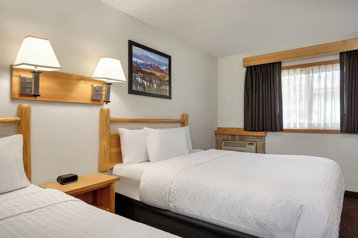 A hotel room with two beds, a wooden headboard, a nightstand, a lamp, a painting, and a window with curtains.