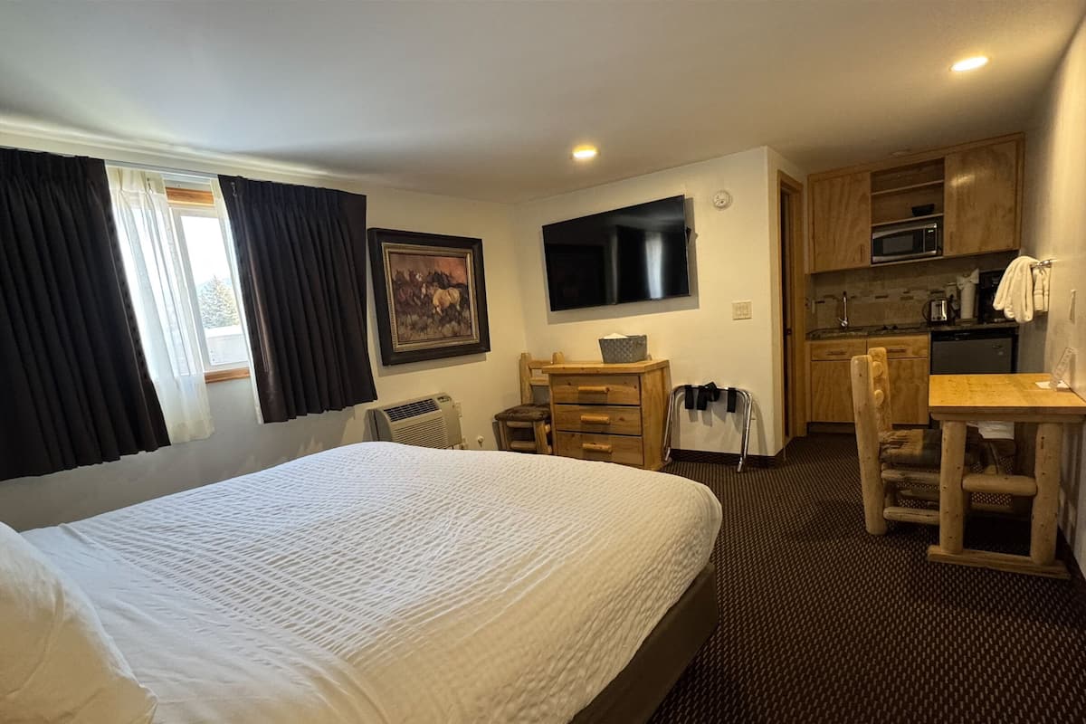 A hotel room with a large bed, a desk, a chair, a television, a painting, a microwave, and a kitchenette.