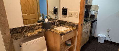 Baño con inodoro, lavamanos y espejo. Hay un armario de madera y un cubo de basura.