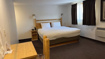 A hotel room with a wooden bed, a small bedside table, a chair, and a window with curtains.