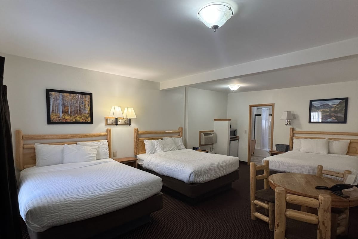 A hotel room with three single beds, a small table, and a chair. There are wall-mounted lamps and a ceiling light.