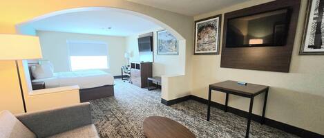Une chambre d’hôtel équipée d’un canapé, d’une petite table, d’un bureau, d’une chaise, d’un lit, d’une télévision et d’œuvres d’art accrochées aux murs.