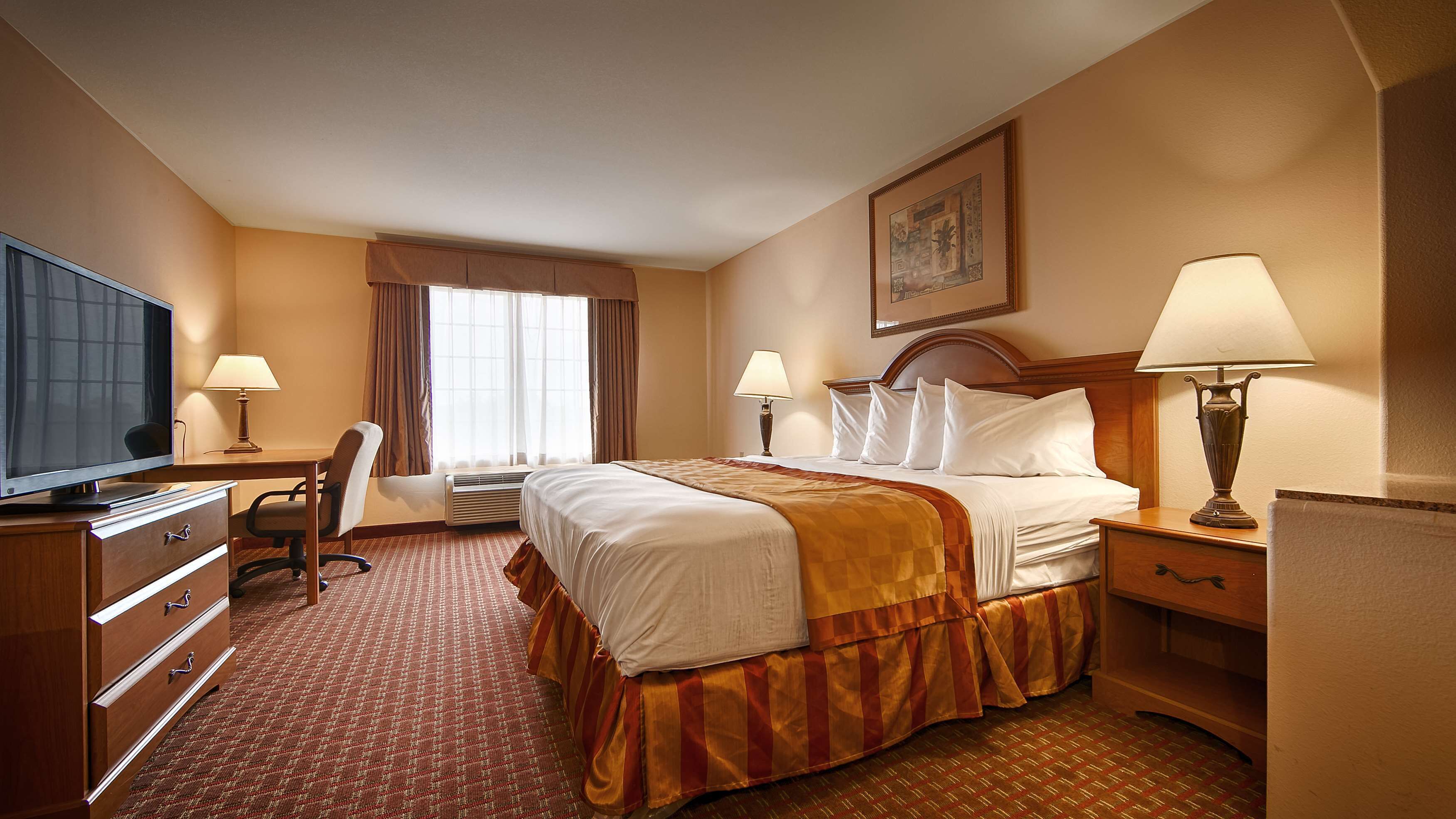 A hotel room with a bed, desk, chair, television, lamps, and a painting on the wall.