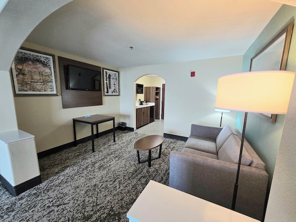 A hotel room with a sofa, a small table, a TV mounted on the wall, and a lamp.