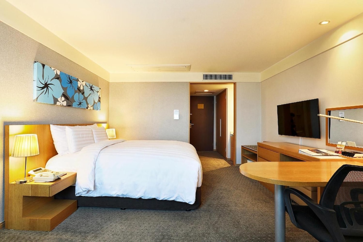 A hotel room with a bed, bedside table, lamp, desk, chair, and a television.
