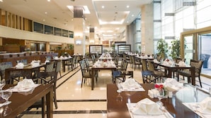 Ein geräumiger Hotel-Speiseraum mit mehreren gedeckten Tischen, einer langen Theke mit Buffetangebot und großen Fenstern für natürliches Licht.