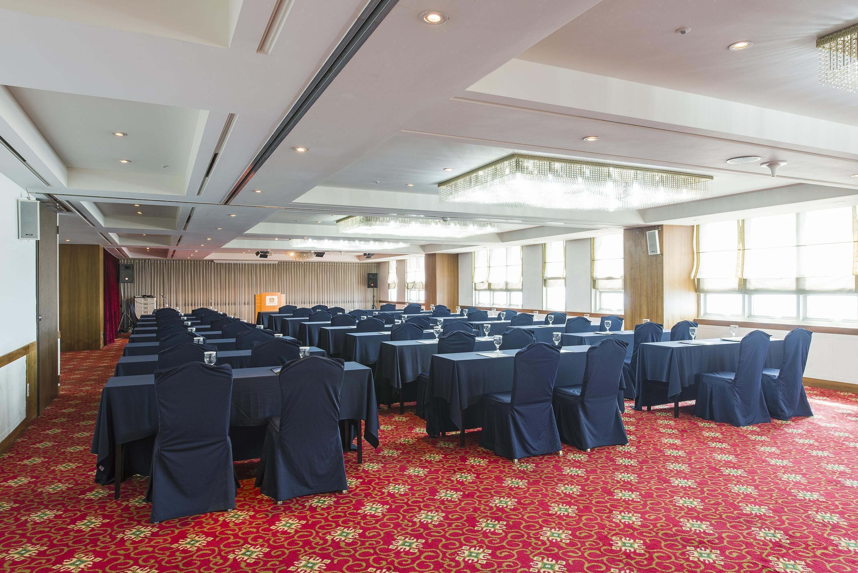 Un salón de conferencias con filas de mesas y sillas, ventanas grandes y una alfombra roja estampada.