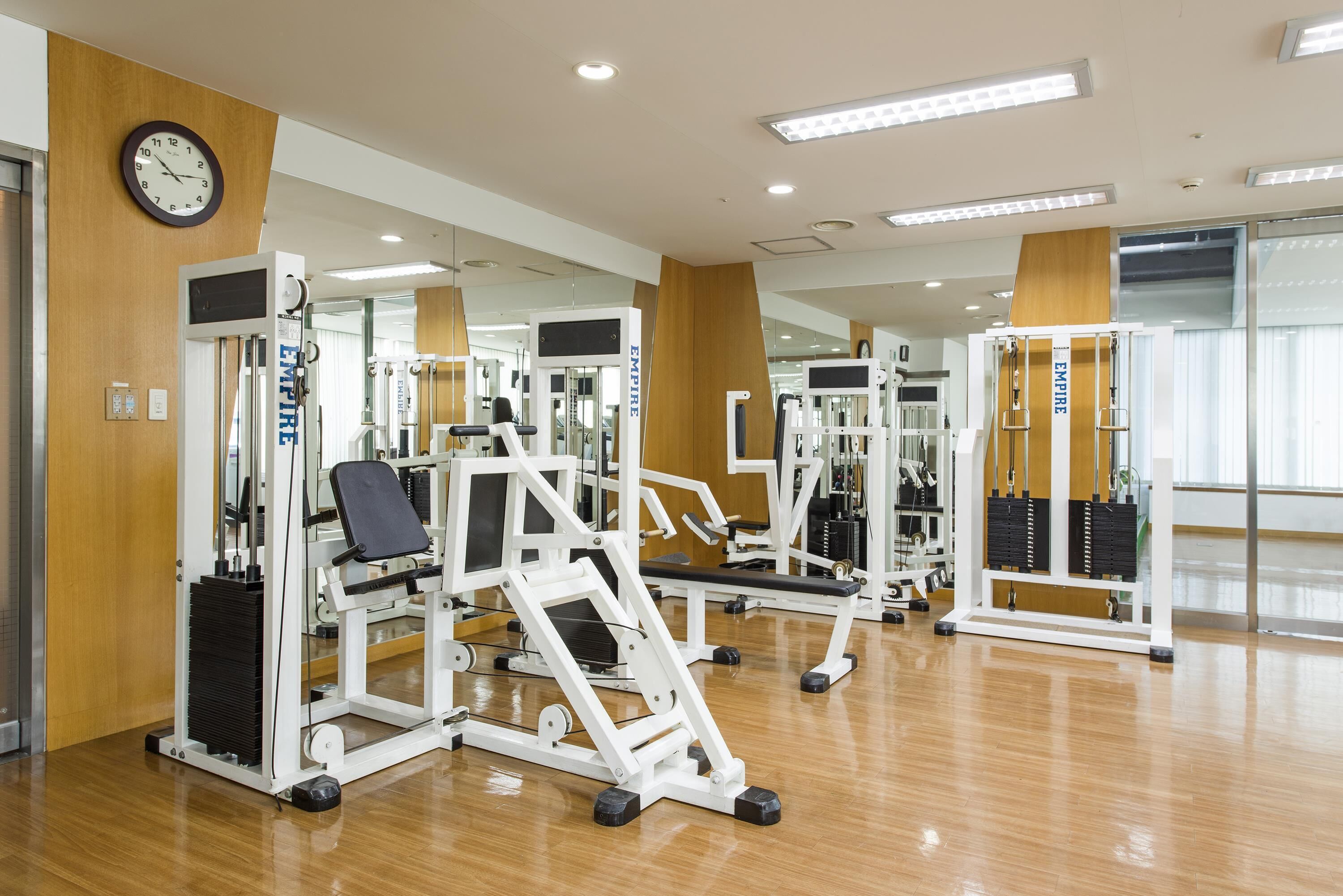 A gym with various exercise machines, a clock on the wall, and a large mirror reflecting the equipment.