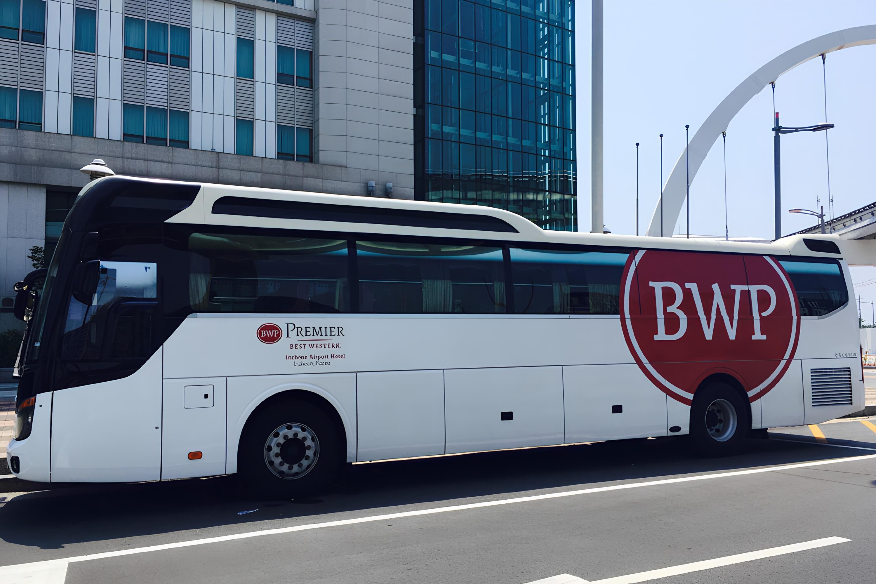 A white bus with 'BWP PREMIER' and 'BEST WESTERN' branding, parked on a road with modern buildings in the background.