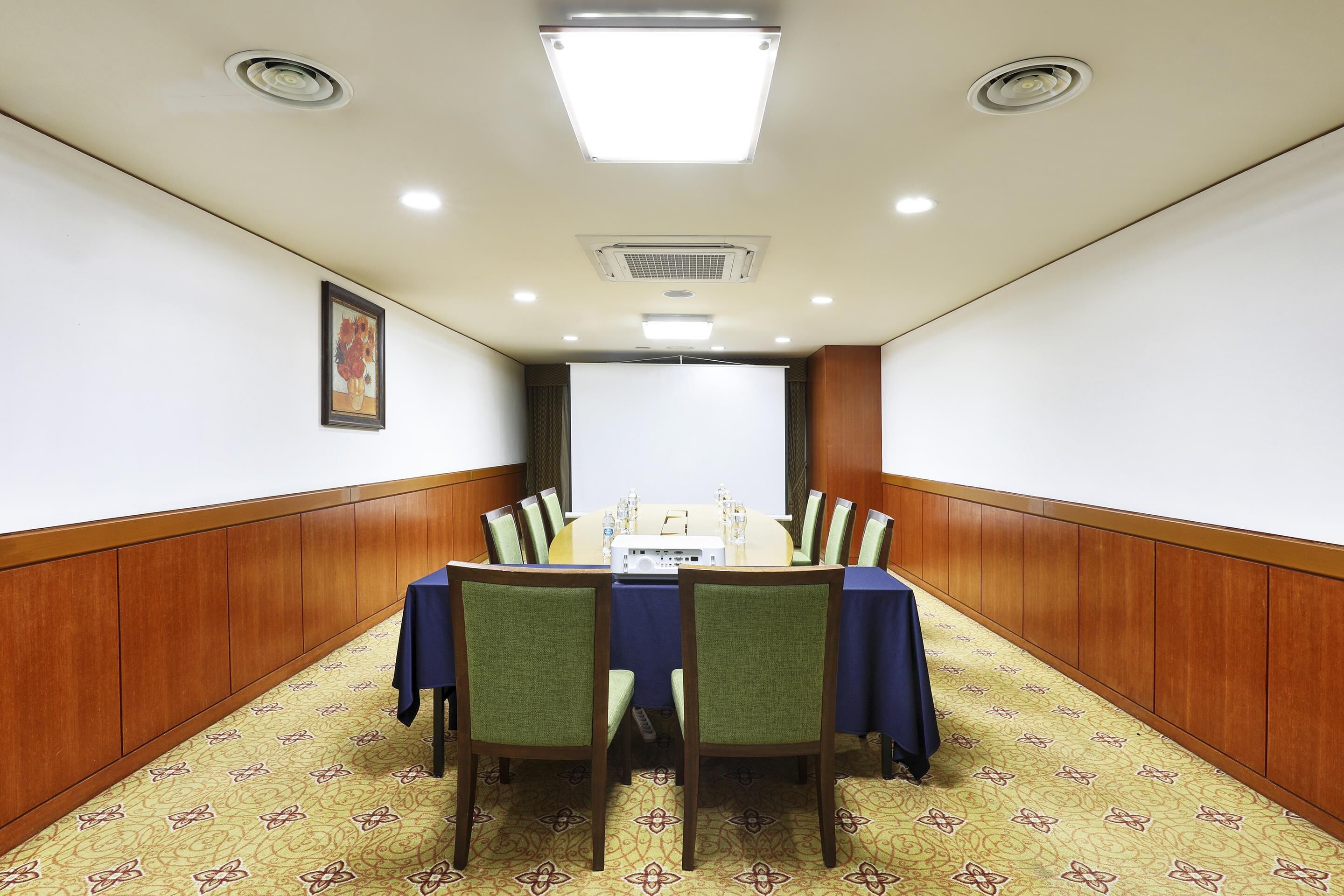 A conference room with a long table, chairs, a projector screen, and a framed picture on the wall.