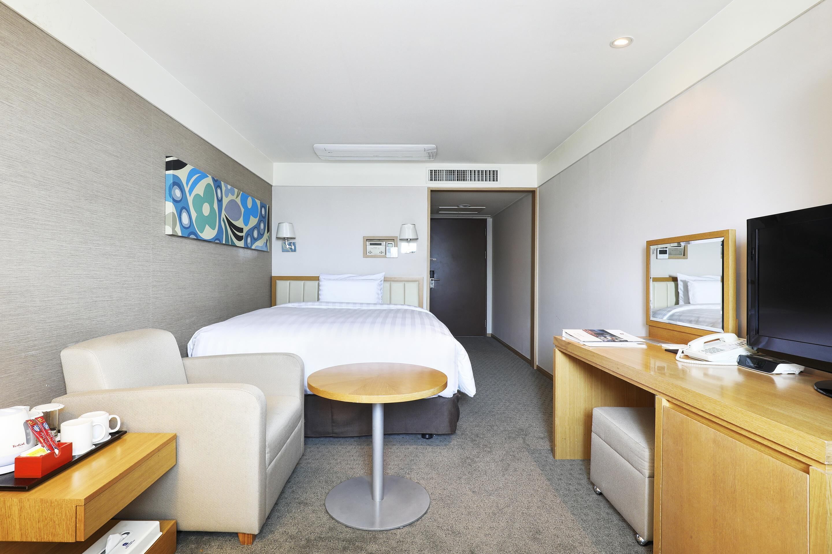 A hotel room with a bed, a desk with a computer, a chair, a small table, and a TV.