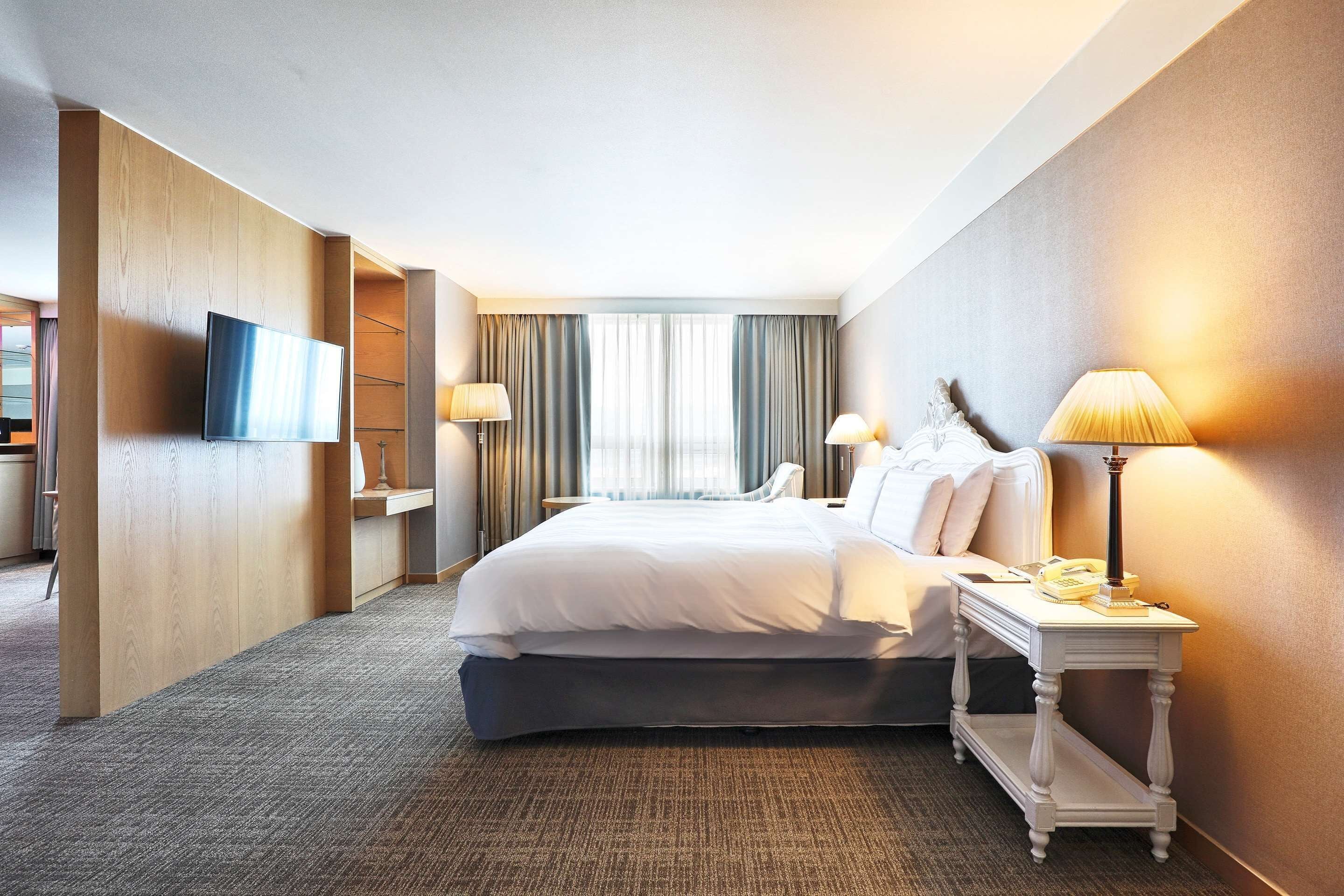 A hotel room with a large bed, two bedside lamps, a flat-screen TV mounted on the wall, and a small table with a telephone and a book.
