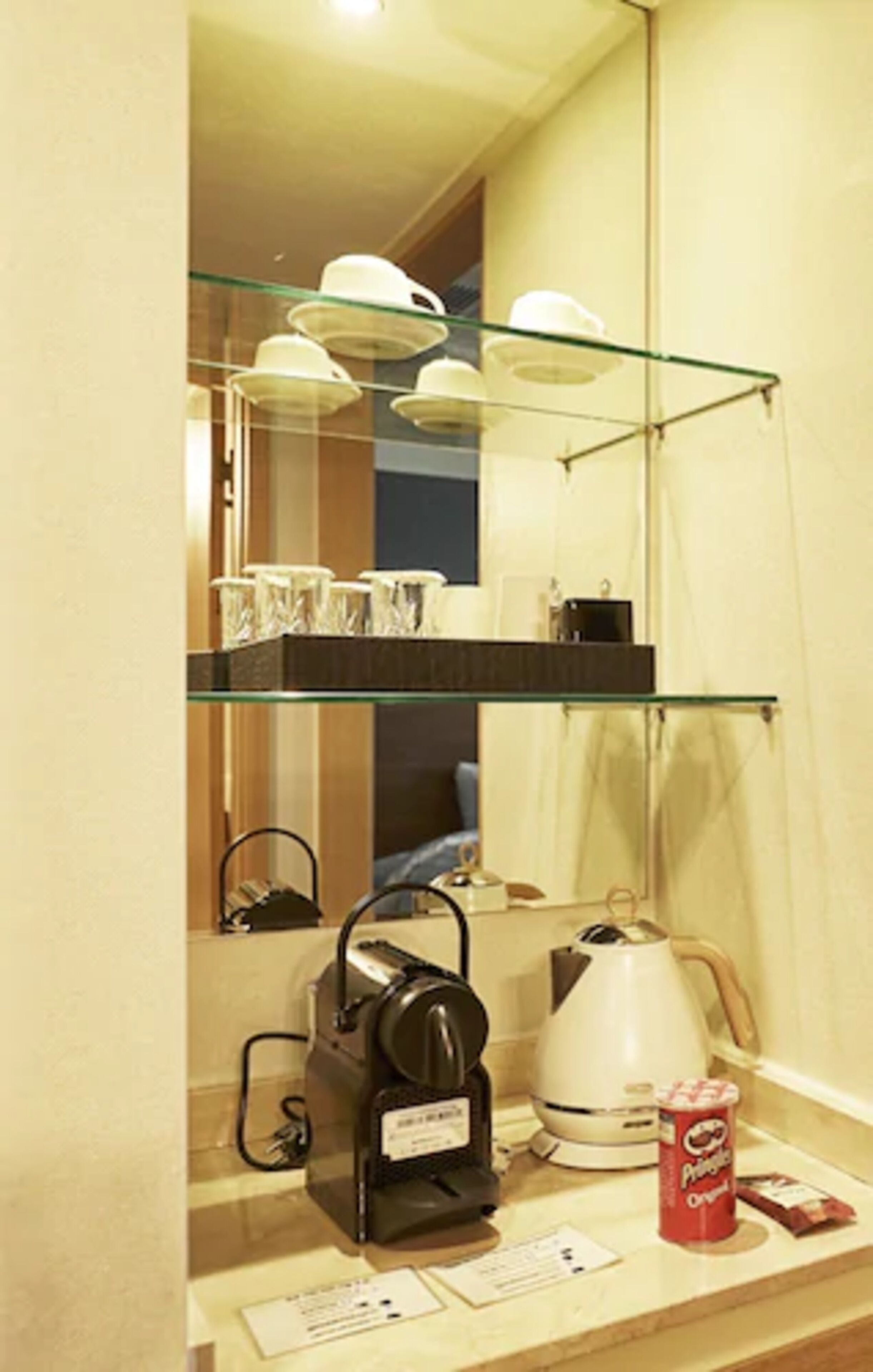 A kitchenette with a coffee maker, kettle, and a glass shelf with cups and a container of Pringles.